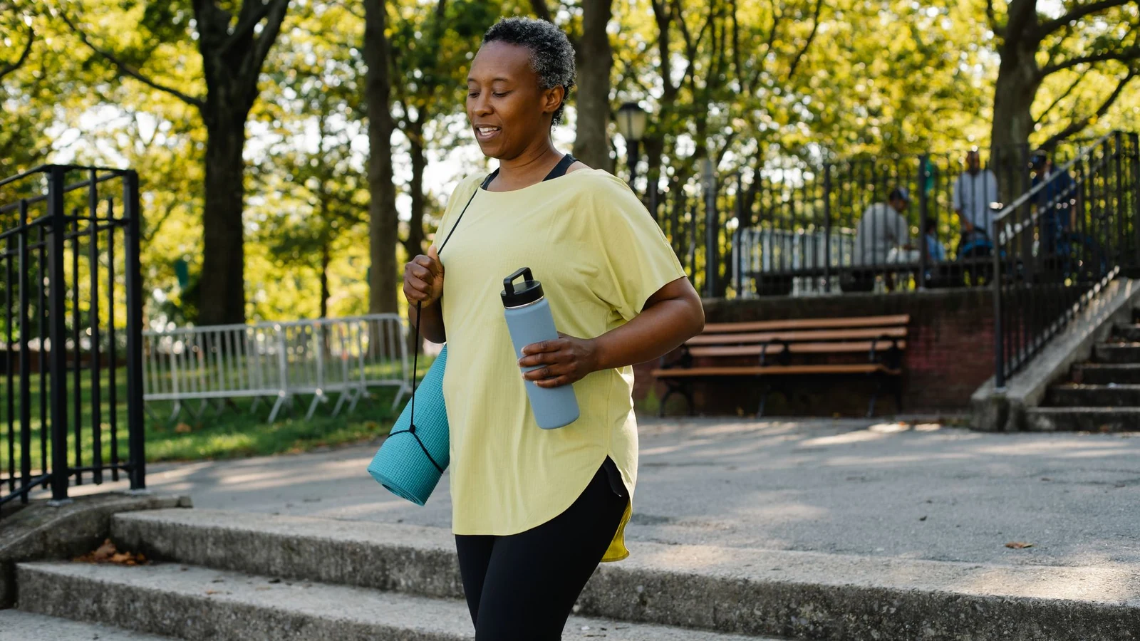 Woman walking outdoors with a water bottle and yoga mat on a sunny day in a public park. — selected for United Kingdom readers