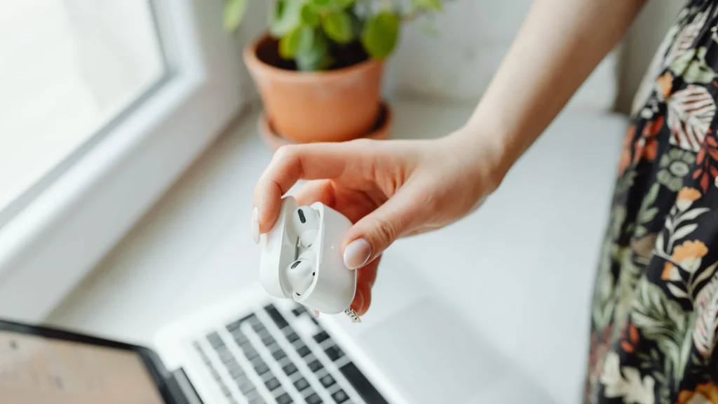 A person holds wireless earbuds over a laptop in a cozy home setting, with natural light from a window. — selected for United States readers