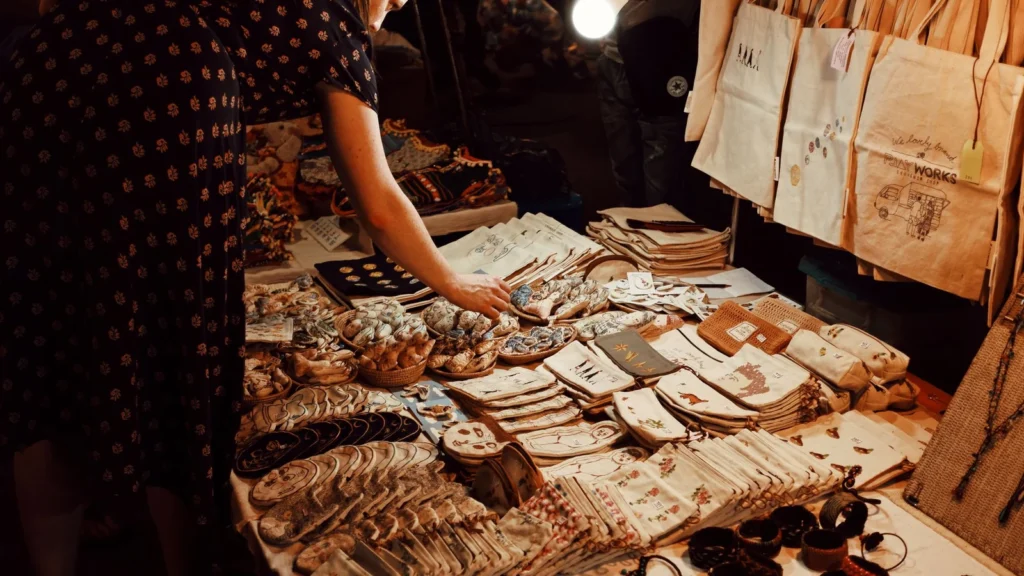 From above crop female customer in dress choosing handmade goods from table in local souvenir bazaar in evening — selected for United Kingdom readers
