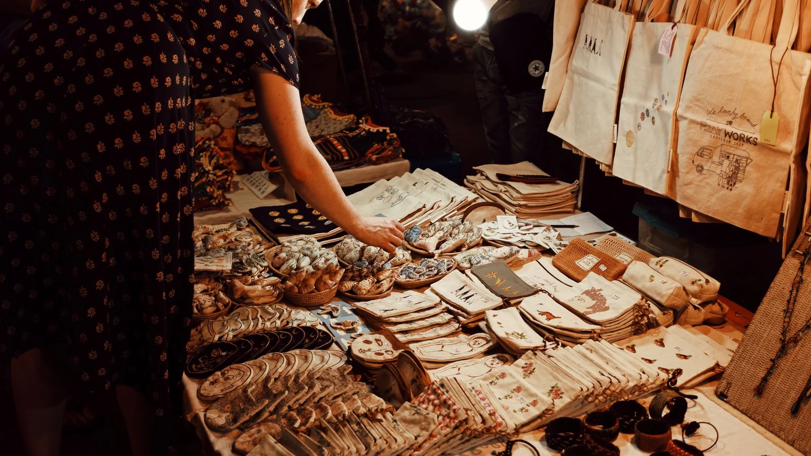 From above crop female customer in dress choosing handmade goods from table in local souvenir bazaar in evening — selected for United Kingdom readers