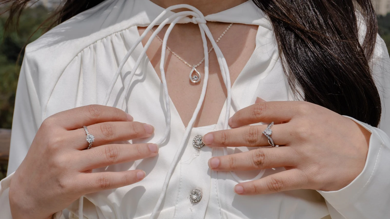 Close-up of a woman wearing elegant jewelry including rings and a necklace with a white blouse outdoors. — selected for United Kingdom readers