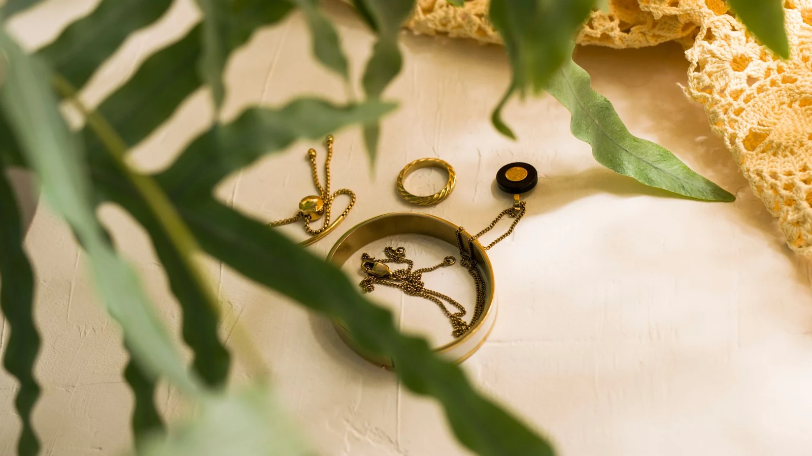 Gold jewelry pieces including bracelets and rings resting on a white textured surface with green leaves and crochet fabric. — selected for United Kingdom readers