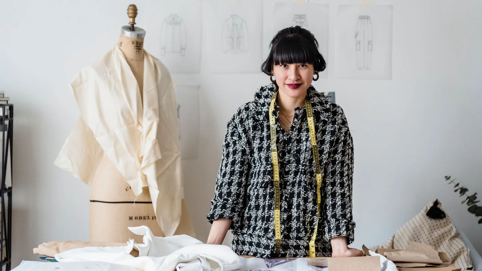 Positive Asian female dressmaker with measuring tape on neck standing at wooden table with fabrics during work in atelier in daytime — selected for United States readers