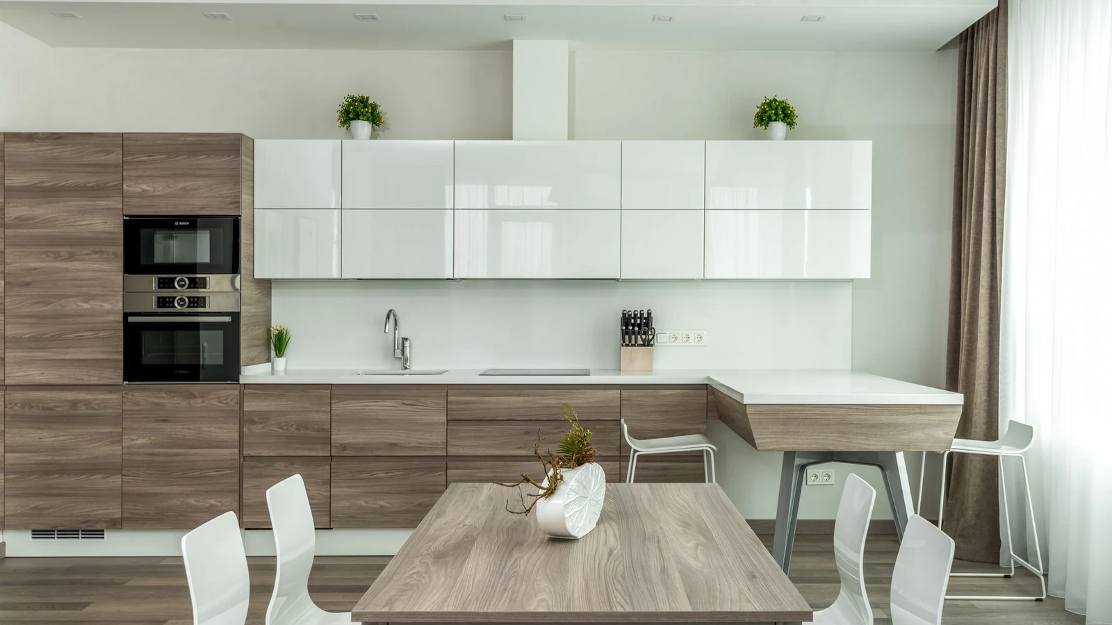 Modern open plan kitchen with white glossy and wooden cabinets and dining table with white chairs near in light apartment — selected for United Kingdom readers