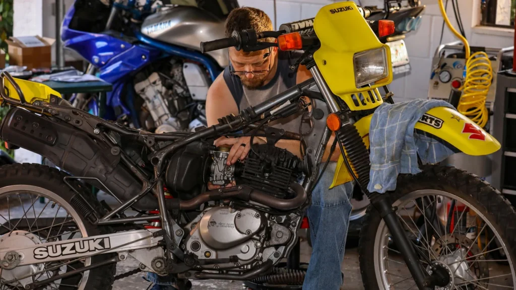 Mechanic repairing a Suzuki motorcycle in a cluttered garage, focusing on engine maintenance. — selected for United States readers