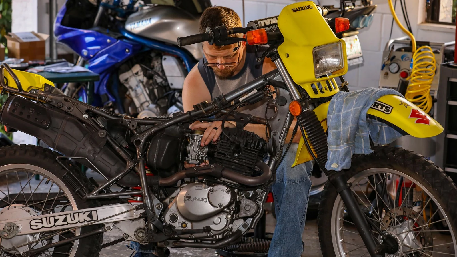 Mechanic repairing a Suzuki motorcycle in a cluttered garage, focusing on engine maintenance. — selected for United States readers