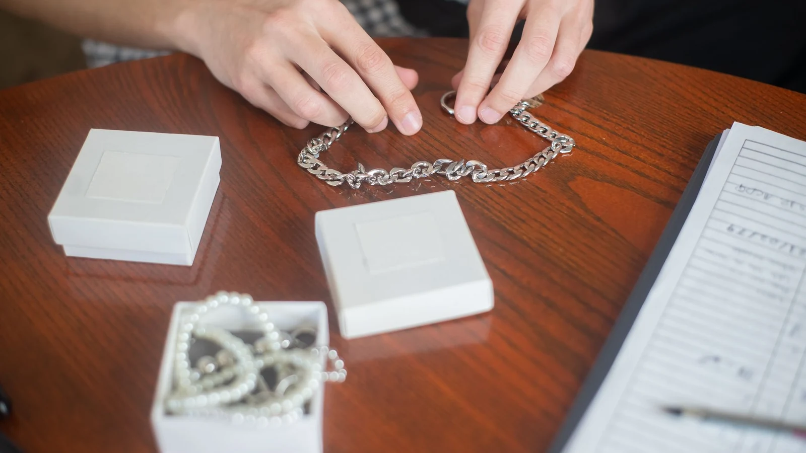 Hands arranging a silver chain necklace on a wooden table with jewelry boxes, indoors. — selected for United Kingdom readers