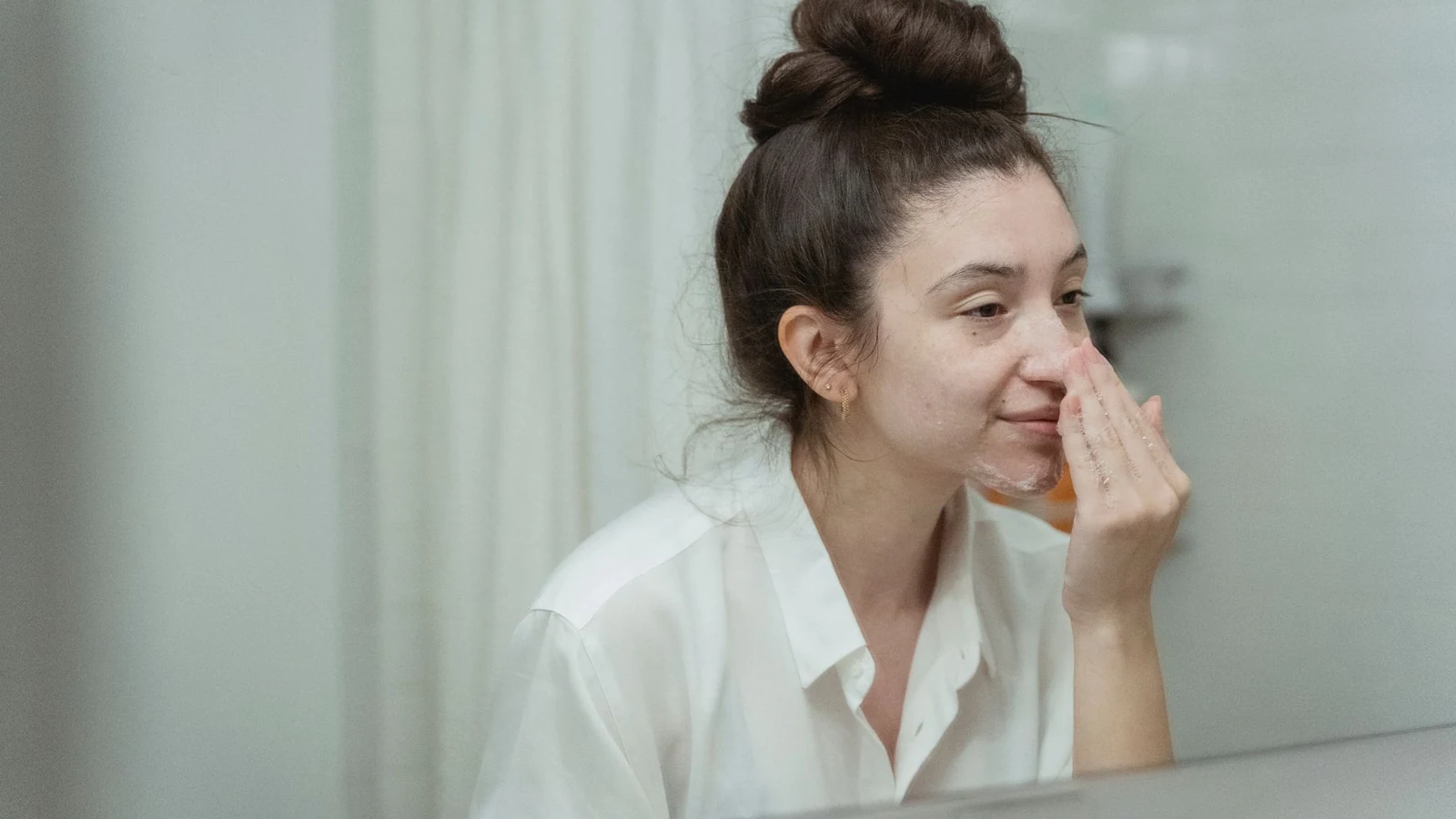 Woman applying face cream in bathroom, focusing on skincare routine with natural reflection in mirror. — selected for United Kingdom readers