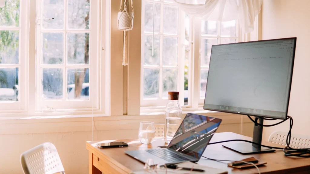 A sunlit home office space featuring a laptop on a wooden desk with natural light streaming through large windows. — selected for United States readers