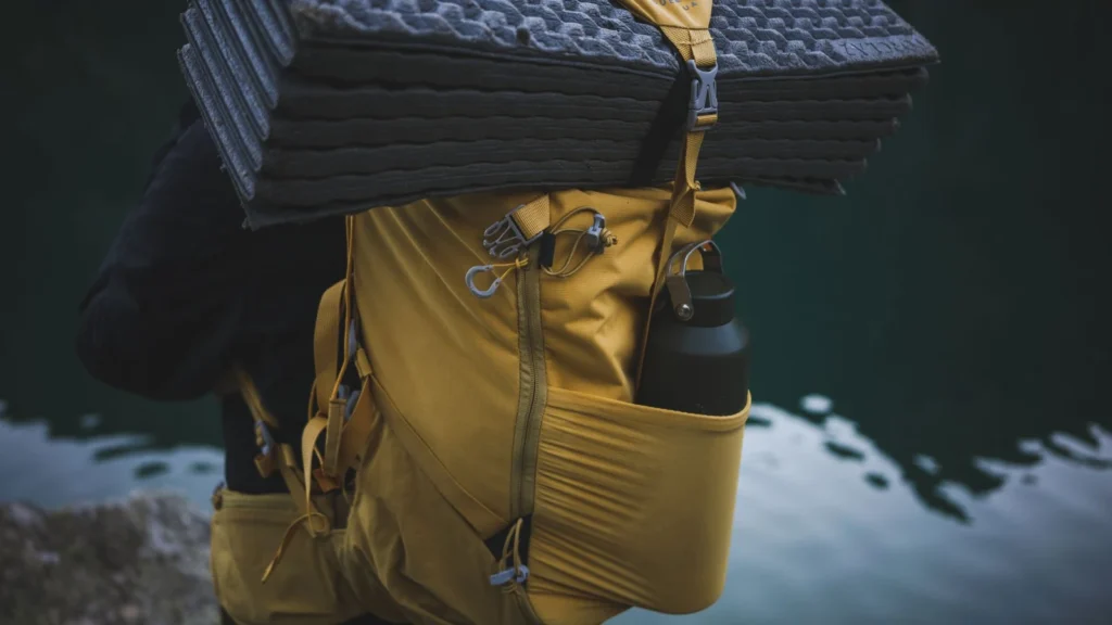 Yellow backpack with camping gear including a water bottle and folded mat near water. — selected for United Kingdom readers