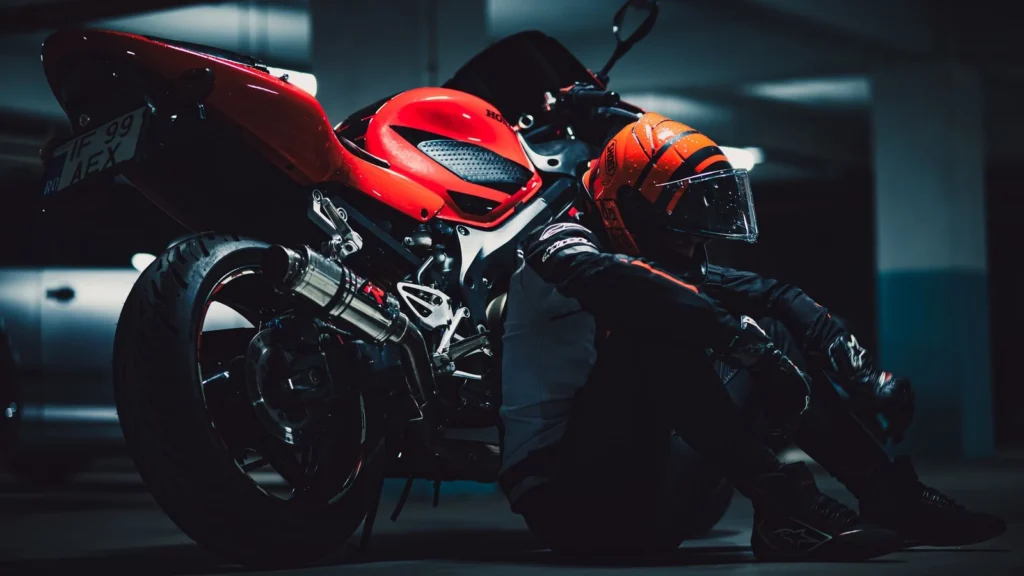 A motorcyclist in full gear relaxes beside a red motorcycle in a dimly lit garage. — selected for United States readers