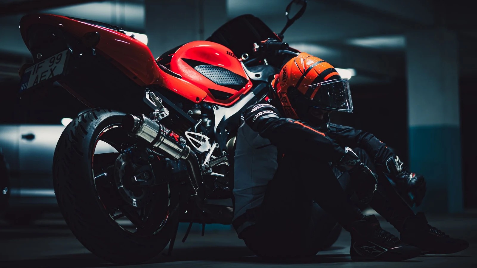A motorcyclist in full gear relaxes beside a red motorcycle in a dimly lit garage. — selected for United States readers